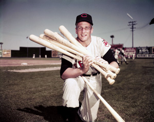 A baseball player in a vintage uniform kneels on a field, smiling and holding several wooden bats. A stadium and other players are visible in the background under a clear sky.