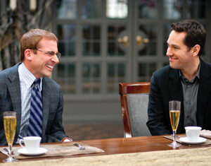 Two men sit at a table, smiling at each other. Both have glasses of champagne and cups in front of them. One wears a suit and tie with glasses; the other wears a dark jacket. The background shows large windows.