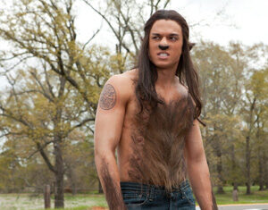 A shirtless person with long hair, fake fangs, a tattoo on their upper arm, and thick, fake fur on their chest and arms stands outdoors near a road, appearing to snarl. Trees with sparse leaves are in the background.