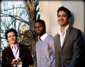 Three men stand outdoors beside a light-colored wall, dressed in collared shirts and jackets. Bare tree branches are visible in the background, and the men are smiling or looking relaxed.