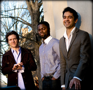 Three men stand outdoors beside a light-colored wall, dressed in collared shirts and jackets. Bare tree branches are visible in the background, and the men are smiling or looking relaxed.