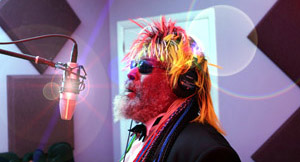 A man with a colorful wig, sunglasses, and headphones sings into a microphone in a recording studio with soundproof panels on the wall and lens flare effects in the image.