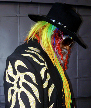 A person in a black hat and sunglasses wears a black jacket with bold gold patterns. Their hair and beard are brightly colored in rainbow shades, featuring braids and dreadlocks. The person is facing downward.