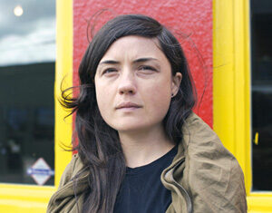 A woman with long dark hair stands in front of a red wall with a yellow window frame, holding a black helmet and wearing an olive green jacket, looking thoughtfully into the distance.