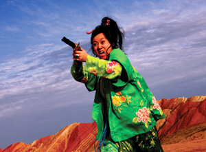 A woman in a vibrant green floral outfit points a gun forward with an intense expression, standing in a colorful, rocky landscape under a cloudy sky.