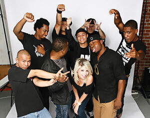 A group of nine people pose playfully in front of a white backdrop, making expressive hand gestures and smiling. One woman is in the center, surrounded by eight men.