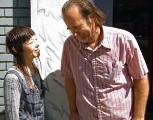 A woman and a man stand outside in sunlight, facing each other and smiling. The woman has dark hair with bangs and wears a patterned shirt; the man has light brown hair and wears a short-sleeve, striped shirt.