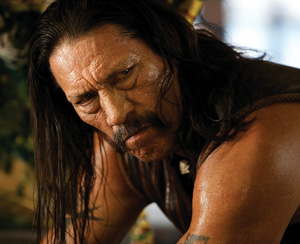 A man with long dark hair and a mustache stares intensely, leaning forward. His muscular arms and sweat on his skin suggest physical exertion. He wears a sleeveless leather vest.