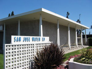 A modern building with angled white pillars and a textured gray facade. In front, a white decorative concrete wall displays blue letters reading San Jose Water Co. Landscaping with plants and flowers surrounds the entrance.