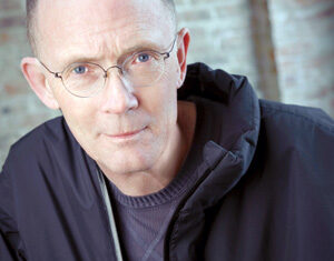 A middle-aged person with glasses, short light hair, and a serious expression wears a dark jacket and sweater, standing in front of a blurred brick wall background.