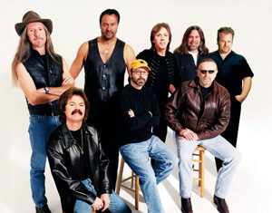 Eight men pose together in a studio with a white background, wearing casual and leather jackets; some are seated on stools while others stand behind them.