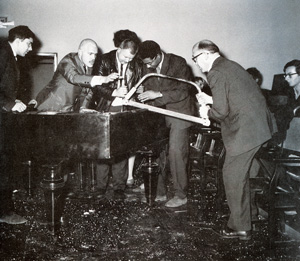 A group of men surround a grand piano, with one man sawing through its frame while others watch and assist. The scene appears intense and takes place in a room with chairs and an audience.