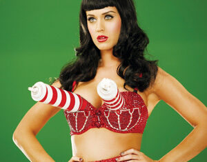 A woman with long dark hair and bangs wears a sequined red bra with large, white and red striped cone shapes attached, standing against a plain green background with her hands on her hips.