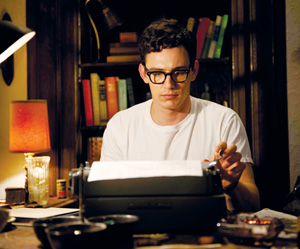 A man wearing glasses and a white t-shirt sits at a desk, typing on a typewriter, surrounded by books and a lit lamp in a cozy, dimly lit room.