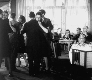 Black and white photo of couples ballroom dancing, while several people sit at tables in the background, eating, drinking, and watching the dancers in a lively, elegant indoor setting.