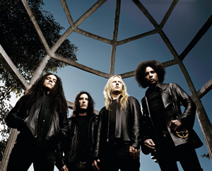 Four men with long hair, dressed in black leather jackets, stand under a geometric metal structure with a blue sky above and trees in the background, looking seriously at the camera.