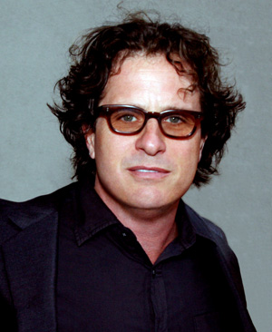 A man with curly dark hair wearing brown glasses and a dark suit jacket over a dark shirt, standing in front of a plain gray background.
