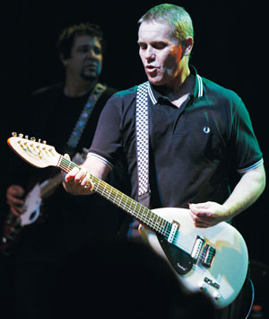 A man in a black polo shirt with a checkered strap plays a white electric guitar on stage, with another musician blurred in the background.