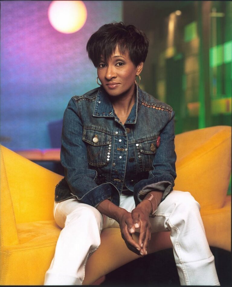 A woman with short dark hair, wearing a denim jacket and white pants, sits on a yellow chair with her hands folded. The background is colorful and softly lit, with shades of blue, green, and purple.
