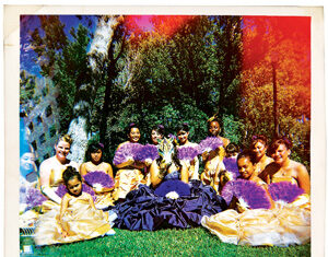 A group of girls and young women in yellow dresses holding purple fans sit on grass outdoors, gathered around a central figure in a dark dress, with trees and sunlight in the background.