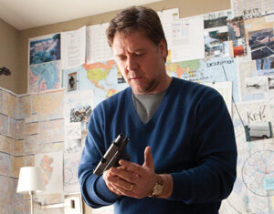 A man in a blue sweater stands in front of a wall covered with maps and papers, holding and inspecting a handgun near an open suitcase.