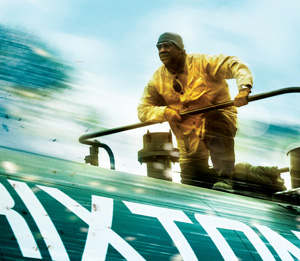 A man in a yellow jacket and dark head covering stands on top of a speeding train, gripping a rail, with blurred motion and the partial word BRIXTON visible on the trains side.