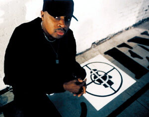 A person wearing a black cap and dark clothing sits next to a painted symbol on the ground, featuring a crosshair design. The setting appears to be indoors with a white brick wall in the background.