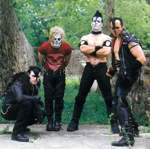 Four men pose outdoors by stone walls, wearing punk and goth-inspired outfits. Two have painted faces with skull designs, and one is shirtless with a mohawk. The scene suggests a dark, alternative music style.