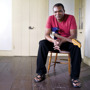 A man in a red shirt and black pants sits on a wooden chair in a simple room, holding an electric guitar across his lap and looking at the camera.