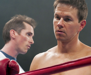 A serious-looking boxer stands in a boxing ring, shirtless, with another man in the background, slightly out of focus, watching him intently.