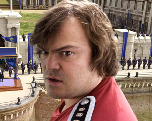 A man with medium-length brown hair, wearing a red shirt with epaulets, looks surprised while standing in front of a large stone building with columns and blue banners. People are gathered in the background.