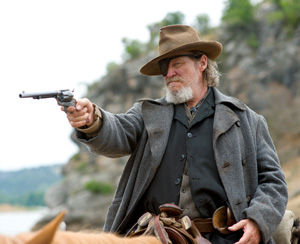 A bearded man wearing a wide-brimmed hat, gray coat, and eye patch sits on a horse, aiming a revolver with his right hand in an outdoor setting with rocky hills in the background.