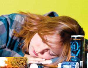 A young woman with red hair sleeps with her head on a desk cluttered with energy drinks and supplement bottles, against a yellow background.