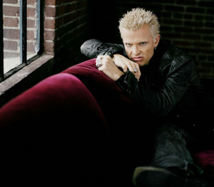 A man with spiky blond hair and a black leather jacket leans on a red velvet couch by a window with exposed brick walls, looking intently at the camera.