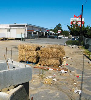A vacant lot with scattered trash, three stacks of hay bales, a concrete structure in the foreground, and a commercial building with signage and a parking lot in the background under a clear blue sky.