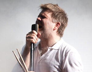 A man wearing a white polo shirt holds drumsticks in one hand and passionately sings into a vintage microphone, with a misty or smoky background.