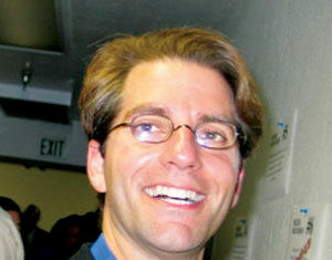 A man with light brown hair and glasses smiles at the camera. He is wearing a blue shirt under a dark jacket and standing indoors near a wall with papers on it. Other people are visible in the background.