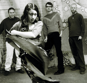 Four people stand in front of a mural. Three men are in the background, and a woman in the foreground is moving dramatically, creating a motion blur with a flowing piece of fabric. The image is in black and white.