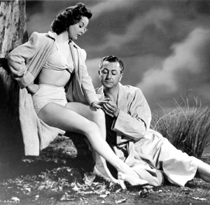 A woman in a bikini top, high-waisted shorts, and an open coat sits beside a man in a robe. He kneels, holding her hand, as they interact in a dramatic outdoor setting with tall grass and cloudy sky.