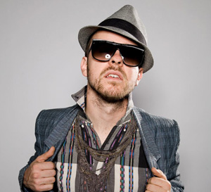 A man wearing a gray fedora, large black sunglasses, a striped shirt, and a gray blazer poses confidently, gripping his jacket lapels.