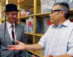 Two men talking in a storage room with shelves of plastic bins. One man wears a suit, tie, hat, and has long braids; the other wears glasses and a light shirt, gesturing with open hands while speaking.