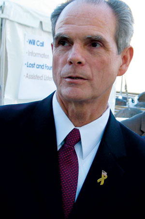 A man in a dark suit and red tie, wearing a gold ribbon pin, stands outdoors in front of a tent with informational signs in the background.