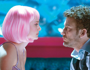 A woman with a short pink wig and a man with curly hair sit closely, facing each other intensely, against a vibrant blue and pink background.