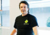 A man with dark hair stands indoors, smiling, wearing a black t-shirt with a green logo and SV Care text on the front. He rests his hands on a glass surface in a well-lit, modern space.