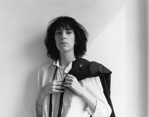 Black-and-white photo of a person with dark, shaggy hair wearing a white shirt and tie, holding a dark jacket over one shoulder, standing against a plain wall and looking confidently at the camera.