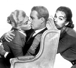 A man and a woman sit closely, about to kiss, while another woman sits behind the chair, looking at them with a serious expression. The scene is in black and white.