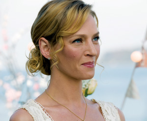 A woman with blonde hair styled in a loose bun, wearing a white lace dress and a gold necklace, smiles softly against a blurred outdoor background with soft lighting.