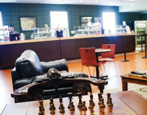 A chessboard set up for a game sits on a wooden table in the foreground of a cozy café, with a leather sofa, chairs, and a counter visible in the background.