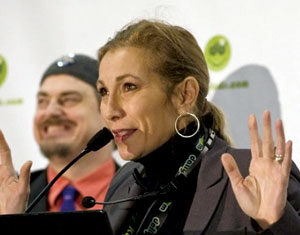 A woman speaks into a microphone at a podium, raising her hands. She wears a dark blazer and hoop earrings. A smiling man in a hat and tie stands in the background. A white banner with green logos is behind them.