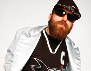 A bearded man wearing dark sunglasses, a black flat-brimmed cap, a shiny silver jacket, and a black hockey jersey with a shark logo poses confidently against a plain white background.